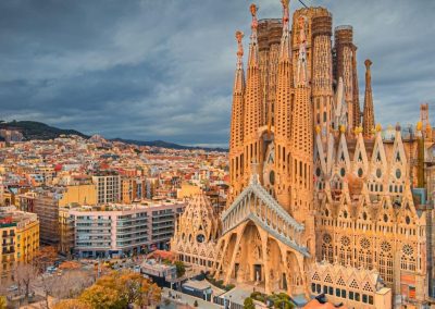 The awe-inspiring Sagrada Família in Barcelona, Spain, an intricate basilica with towering spires against a dramatic cloudy sky, overlooking the colorful cityscape.
